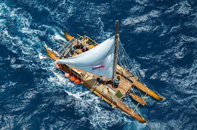 Double-hulled canoe viewed from overhead while it sails. The sail is filled with wind and crew members work on board.